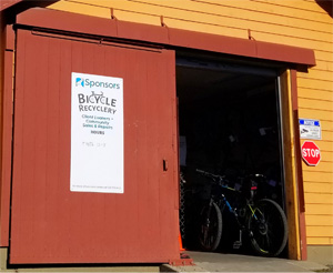 Exterior of yellow and brown building. Barn door is open, revealing a bicycle repair shop. Exterior sign reads 