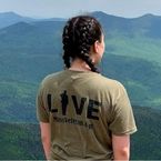 a woman stands on a mountain top, facing the view, with the back of her tshirt reading 