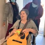 Shayna, who is wearing a hospital gown and holding her guitar, smiles as she sees her new home for the first time.