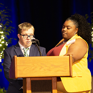 A man with Down syndrome speaks at a podium with the assistance of his direct support professional