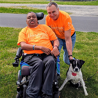 Two men posing side by side in orange shirts, one man is in a wheel chair. A black and white dog is being held by the other man.