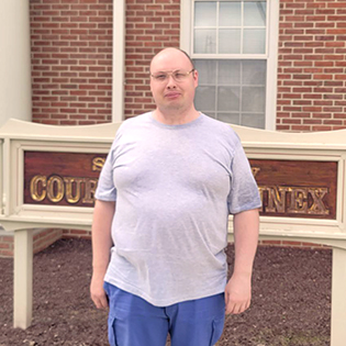 man with a disability stands in front of Sussex County Court sign. He is wearing a gray t-shirt  and glasses. He is smiling at the camera. 