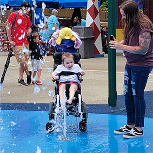photo of a girl in a wheelchair enjoying a splash pad at Holiday World while a woman takes a photo using her phone