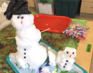 Two snowmen stand on a tray in a classroom. One wears a hat.