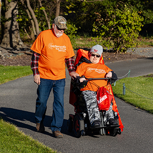 a man in a wheel chair and his father strolling along a cement path in bright orange shirts