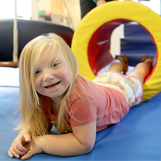 A child with blonde hair and a pink shirt lays on a blue mat and smiles at the camera