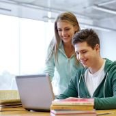 A male high school student wearing a green hoodie uses a laptop with a female classmate wearing a light blue shirt looks on.